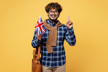 Young teen Indian boy IT student he wear casual clothes shirt glasses bag hold cover half of face with British flag point finger up isolated on plain yellow background. High school university concept.