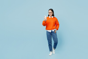 Full body young fun woman of Asian ethnicity wear orange sweater glasses hold use mobile cell phone...