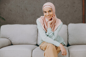 Young smling fun happy calm muslim woman wear hijab casual clothes sits on sofa couch stay at home flat rest relax spend free spare time in living room indoor. People uae middle eastern islam concept.