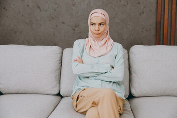 Young sad mad muslim woman wears hijab casual clothes look aside sits on sofa couch stay at home flat rest relax spend free spare time in living room indoor. People uae middle eastern islam concept.
