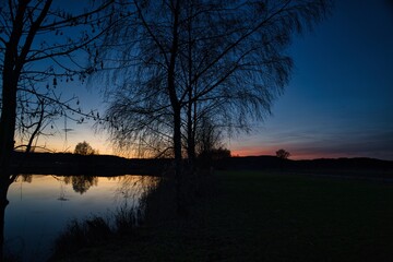 Fototapeta premium Wonderful blue golden red sky after sunset with trees in foreground at a lake with its reflections 