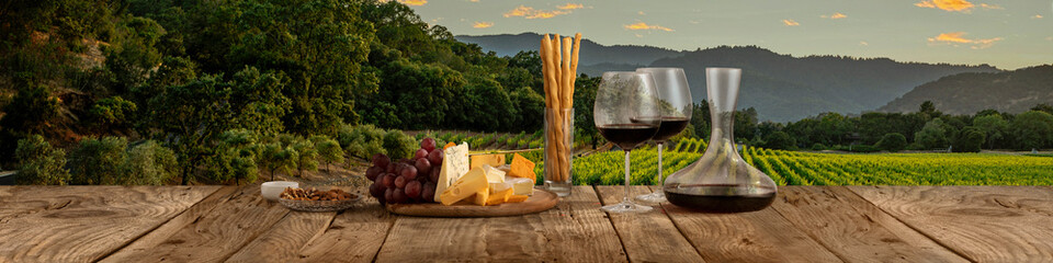 Decanter and two glasses with delicious red wine, grape and cheese appetizers standing on wooden table over beautiful nature landscape of fields and forest.