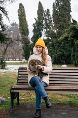 Fototapeta premium A young woman in a yellow hat is holding and stroking a spotted cat while sitting on a bench in nature in the middle of winter