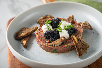 Tatar roh vom Rind mit schwarzen Kaviar, Schmand, Dill und Brot als Gourmet Rindertatar auf Steingut Teller, grün Serviette, Holzbrett und Marmor Hintergrund hell