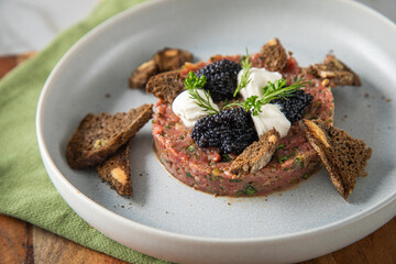 Tatar roh vom Rind mit schwarzen Kaviar, Schmand, Dill und Brot als Gourmet Rindertatar auf Steingut Teller, grün Serviette, Holzbrett und Marmor Hintergrund hell
