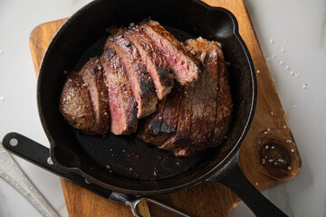 Entrecote Steak vom Rind medium rare gebraten in Gusseisen Pfanne mit Meersalz auf Holzbrett mit Messer, Fleischgabel und hellen Marmor Hintergrund