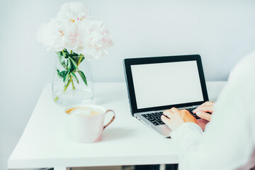 No face woman working at home using a laptop with blank screen for mockup. Cozy workspace with fresh pink peonies flowers and a cup coffee on the white table. Remote work at home. Copy space.