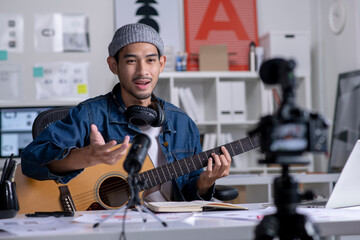 Young Asian male graphic designer blogger influencer playing guitar talking looking at camera while shooting education tutorial vlog training filming video course for social media at studio. - 558675905
