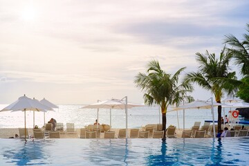White sand beach and blue sea landscape view of a tropical resort hotel beside modern swimming pool, white parasols, palm trees, deck beach chairs in the golden sunset time