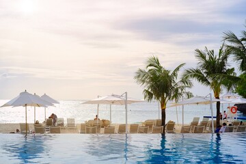 White sand beach and blue sea landscape view of a tropical resort hotel beside modern swimming pool, white parasols, palm trees, deck beach chairs in the golden sunset time