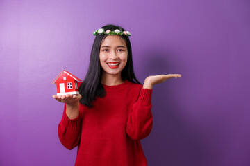 Portrait of beautiful trendy asia cheerful girl holding home toy model showing copy space ad isolated over violet color background, for valentine day color tone