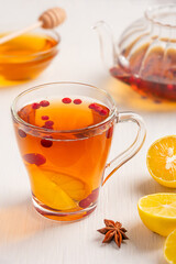 Homemade organic herbal hot tea made with sour sliced lemon citrus, red cranberries and sweet honey served in transparent glass cup on white wooden table with ingredients and teapot. Vertical image
