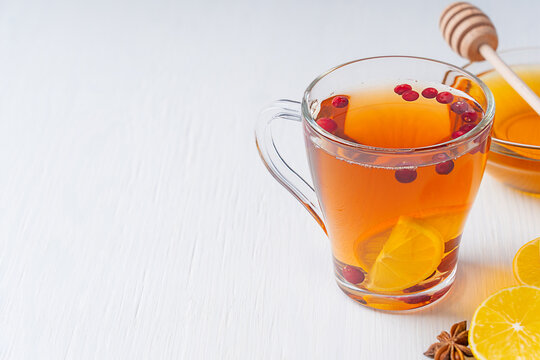 Natural herbal spiced hot tea made with sour sliced lemon citrus, cranberries and sweet honey served in transparent glass cup on white wooden table with ingredients and anise. Image with copy space