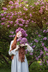 Obraz premium young girl with long red hair with a wreath of lilacs on her head in the garden in spring