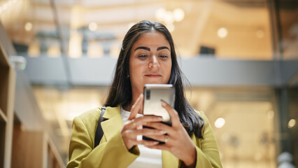 Portrait of a Young Hispanic Woman Using Her Smartphone, Walking in a Busy Office Hall and Smiling. Medium Shot. A Businesswoman in Smart Casual Clothes Enters her Workplace in the Morning