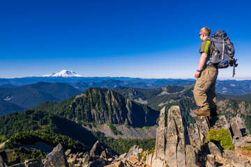 Athletic adventurous male hiker standing on top of a mountain with Mount Rainier in the background during a beautiful sunrise. 
