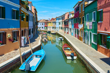 Fototapeta na wymiar Panoramic view of canal with boats in Burano, Italy, surrounded by picturesque colorful houses decorated with plants, flowers and hanging clothes under blue sky on sunny spring day.
