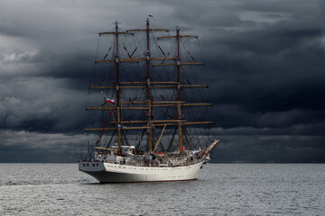Fototapeta na wymiar Three-masted sailing frigate goes to sea from the port against a stormy sky.