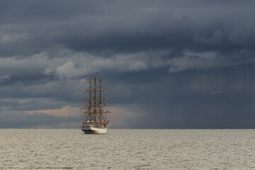 Three-masted sailing frigate goes to sea from the port against a stormy sky.