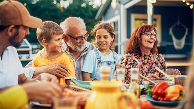 Portrait of a Happy Senior Grandfather Holding His Bright Talented Little Grandchildren on Lap at a Outdoors Dinner Party with Food and Drinks. Family Having a Picnic Together with Children.