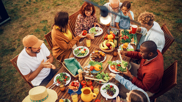 Top Down Elevated View at a Family and Friends Celebrating Outside at Home. Diverse Group of Children, Adults and Seniors Sitting at a Table, Having Fun Conversations. Eating Barbecue and Vegetables.