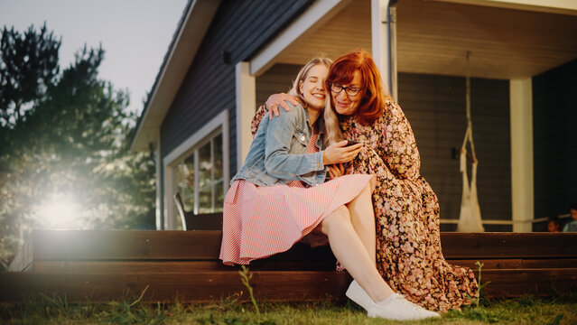 Beautiful Teenage Granddaughter Hugging with Her Grandmother While Using a Smartphone. Girl Showing Family Photos and Videos to Her Grandparent. Relatives Sitting Outside on a Porch on a Warm Day.