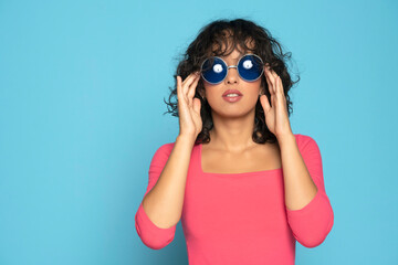 Young exotic brunette woman in pink blouse posing with a sunglasses on a blue background
