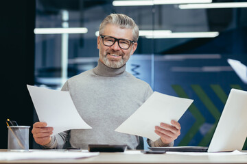 Successful businessman with documents smiling and looking at camera, portrait of mature gray-haired investor financier inside modern office, man behind paper work with reports accounts and contracts