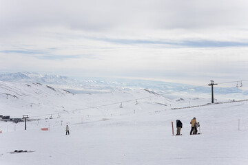 Skiers go downhill skiing on snow-covered mountain ski slope against the backdrop of a cloudy sky and ski lifts. Winter. Extreme sport and travel content  