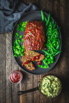 Meatloaf and slices on platter with fresh snap peas, mashed potato in pan, and sauce in jar