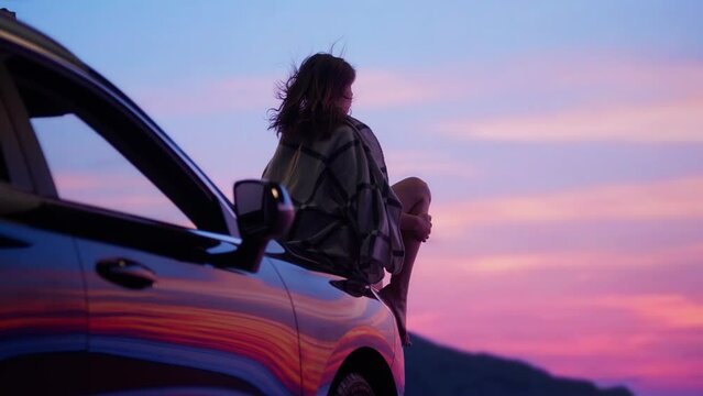 Portrait of beautiful Caucasian female sitting on a car hood, enjoying the view of sunset on a beach