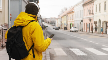 Man in travel yellow jacket and headphones standing on roadside in city holding smart phone in his hand and waiting taxi.