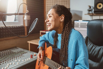 Pleasant cute multiethnic woman singing into microphone and playing guitar in the broadcasting of...