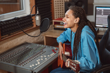 Enthusiastic African young woman musician plays guitar and records song, sings into microphone in...