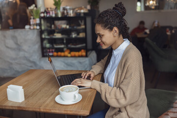 Stylish ethnic student blogger works on laptop in coffee shop. Internet online education, freelance, remote work concept