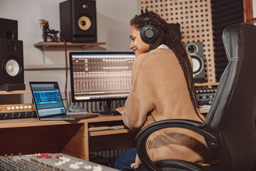 African cute woman blogger records online podcast using laptop, headphones and microphone in professional music studio