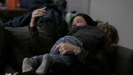 Fototapeta premium Baby climbing on top of mother lying down in sofa and holding smartphone
