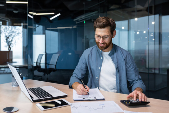 Successful financier investor works with documents and accounts inside office, businessman with beard smiling satisfied with work results and achievements, man inside office with laptop working.