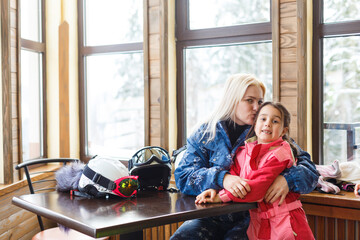 Cheerful mother with daughter on skiing sitting in cafe on ski terrain.