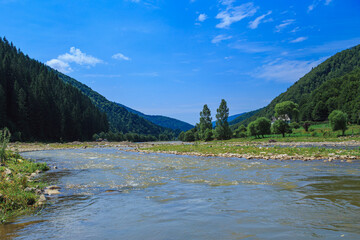 Naklejka na ściany i meble The riverbed of a mountain river with rocky banks. River in a rural mountain area on a sunny day, mountain slopes covered with trees. Sharpness in the center 