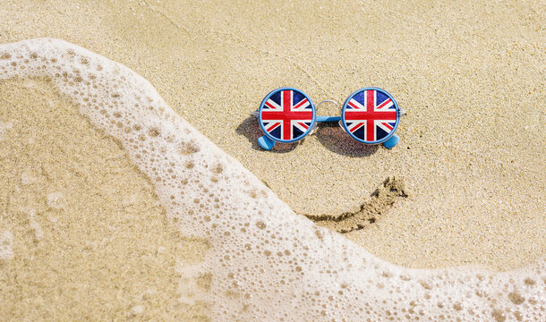 Sunglasses with flag of Great Britain on a sandy beach. Nearby is a sea lightning and a painted smile. Travel and vacation concept for Britons
