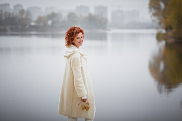 Attractive stylish woman dressed in white warm coat holding yellow leaves walking near river. Autumn mood concept