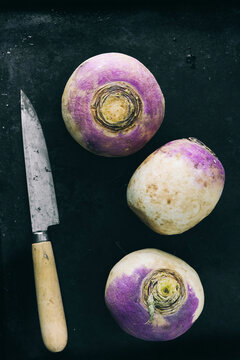 Three purple and white raw turnips with a knife on a black background