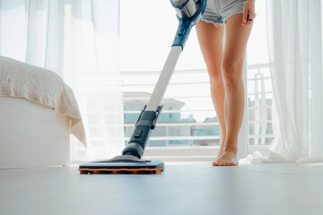 Young woman vacuuming the floor in bright cozy room with cordless vacuum cleaner