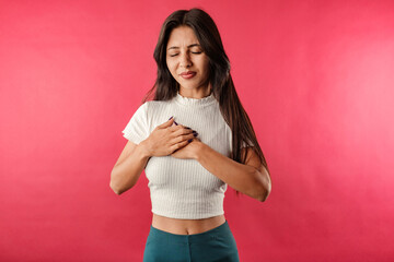 Young beautiful woman wearing ribbed crop isolated over red background having severe chest pain as heart attack and illness concept. Hold hand on chest heart crying posing.