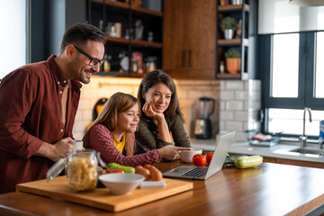 Dad, mother and daughter watching an online cooking class at the kitchen counter at home. Enjoying their time together, cheerfully smiling surrounded with fresh organic food.