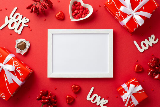 Valentine's Day concept. Top view photo of white photo frame present boxes heart shaped saucer with sprinkles chocolate candies and inscriptions love you on isolated red background with copyspace