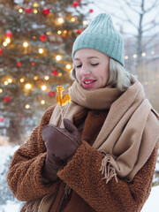 Young smiling woman wearing brown fur coat and hat having fun on a Christmas market and eating rooster-shaped lollipop. Happy beautiful woman having fun on Christmas market. Winter holidays concept.