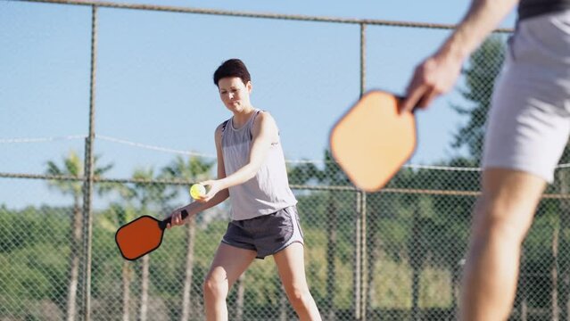 couple playing pickleball game, hitting pickleball yellow ball with paddle, outdoor sport leisure activity.