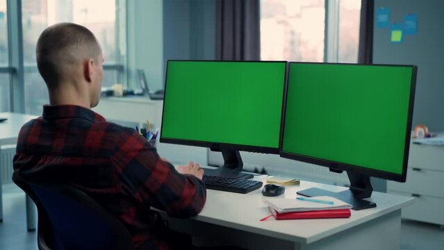 Young Man Working On Computer With Two Green Screen Mock Ups Sitting At Desk In Office